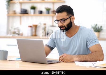 Focused young adult Indian man with eeyeglasses, freelancer, designer or IT specialist, concentrated works from home using laptop, browsing e-mail, internet, is analyzes information Stock Photo