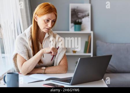 Portrait of adult red haired woman using laptop while working at home office, copy space Stock Photo