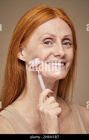 Vertical portrait of beautiful red haired woman with freckles smiling and doing face massage using roller Stock Photo