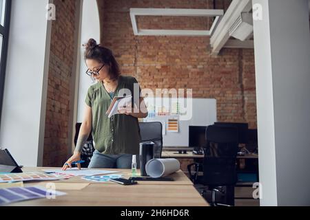 Young woman, interior designer or architect in casual wear with messy hairdo making notes on a blueprint, working on new project, standing in her Stock Photo
