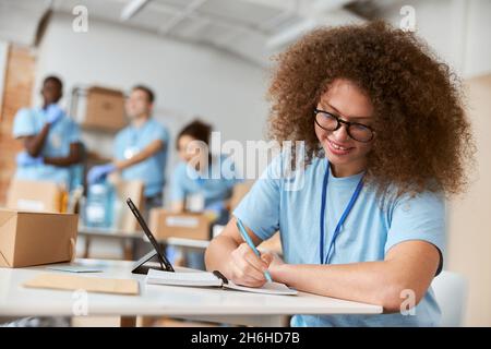 Portrait of young female volunteer in blue uniform making notes while working on donation project. Team sorting, packing items in the background Stock Photo