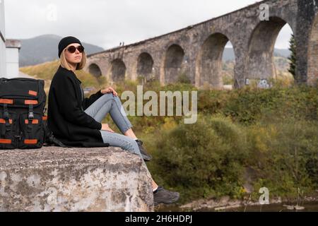 Traveler girl looks at the arched old stone bridge, sitting opposite, the lifestyle of a tourist, traveling across the bridges. Stock Photo