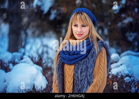 Christmas, New Year holidays, winter concept: happy smiling woman posing in winter park. Model wearing grey faux fur coat, beige sweater and blue scarf and beanie hat. Copy, empty space for text. Stock Photo