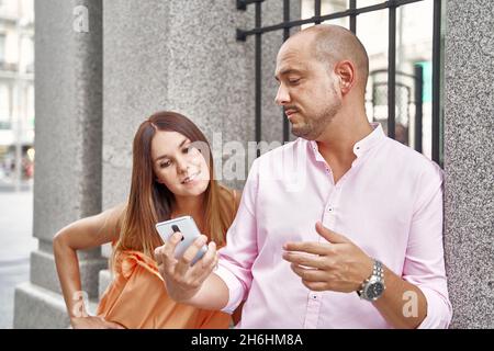 Content stylish boyfriend sharing mobile phone with girlfriend while standing near building in city in summer Stock Photo