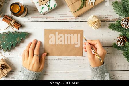 Woman hands in sweater writing on blank envelop greeting card . Flat lay of white wooden background with eco natural Christmas decoration, candle, fir Stock Photo