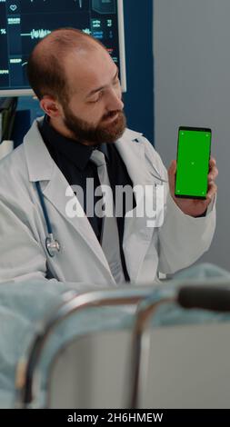 Doctor and sick patient looking at green screen on phone at nursing facility. Medical specialist vertically holding smartphone with chroma key for isolated template and mockup background Stock Photo