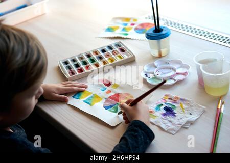 Boy painting pictures with watercolor paints during art lesson. Pupil on drawing with brush. Watercolor color wheel and palette. Color theory beginner Stock Photo