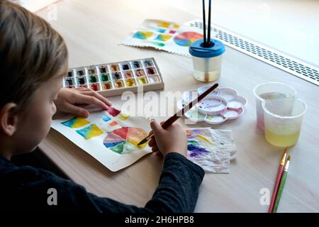 Boy painting pictures with watercolor paints during art lesson. Pupil on drawing with brush. Watercolor color wheel and palette. Color theory beginner Stock Photo
