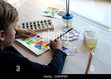 Boy painting pictures with watercolor paints during art lesson. Pupil on drawing with brush. Watercolor color wheel and palette. Color theory beginner Stock Photo