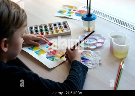 Boy painting pictures with watercolor paints during art lesson. Pupil on drawing with brush. Watercolor color wheel and palette. Color theory beginner Stock Photo