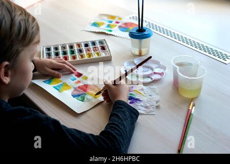 Boy painting pictures with watercolor paints during art lesson. Pupil on drawing with brush. Watercolor color wheel and palette. Color theory beginner Stock Photo