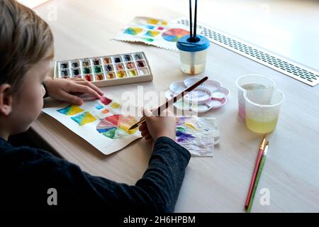 Boy painting pictures with watercolor paints during art lesson. Pupil on drawing with brush. Watercolor color wheel and palette. Color theory beginner Stock Photo