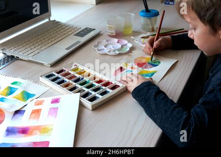 Boy painting pictures with watercolor paints during art lesson. Pupil on drawing with brush. Watercolor color wheel and palette. Color theory beginner Stock Photo