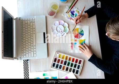 Boy painting pictures with watercolor paints during art lesson. Pupil on drawing with brush. Watercolor color wheel and palette. Color theory beginner Stock Photo