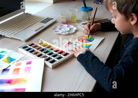 Boy painting pictures with watercolor paints during art lesson. Pupil on drawing with brush. Watercolor color wheel and palette. Color theory beginner Stock Photo