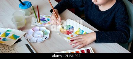 Boy painting pictures with watercolor paints during art lesson. Pupil on drawing with brush. Watercolor color wheel and palette. Color theory beginner Stock Photo