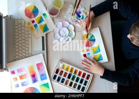 Boy painting pictures with watercolor paints during art lesson. Pupil on drawing with brush. Watercolor color wheel and palette. Color theory beginner Stock Photo