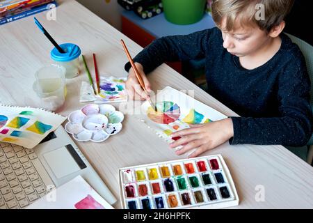 Boy painting pictures with watercolor paints during art lesson. Pupil on drawing with brush. Watercolor color wheel and palette. Color theory beginner Stock Photo