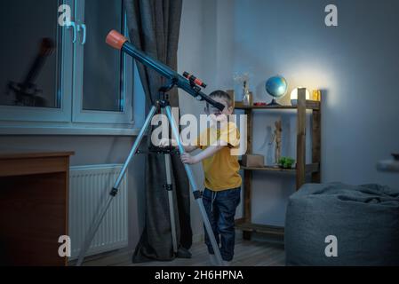 Cute boy is looking through a telescope in a room at the night starry sky. Children's scientific hobbies and space exploration Stock Photo