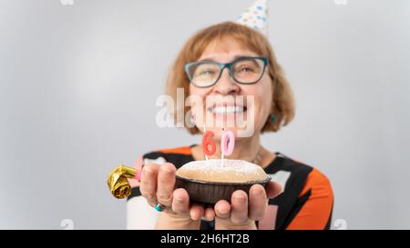 A portrait of a smiling elderly woman in a festive cap holding a cake with candles in the form of the number 60. Anniversary Stock Photo