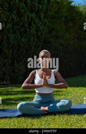 Tranquil African American female with short hair sitting in Lotus pose with prayer hands and closed eyes while doing yoga on mat on lawn in summer Stock Photo