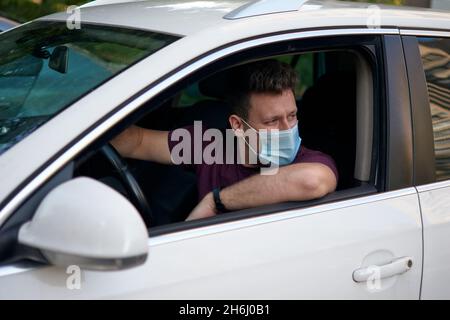 A man driving a car in a medical face mask during coronavirus outbreak, a taxi driver in a mask, protection from the virus. Driver in white car, hand Stock Photo