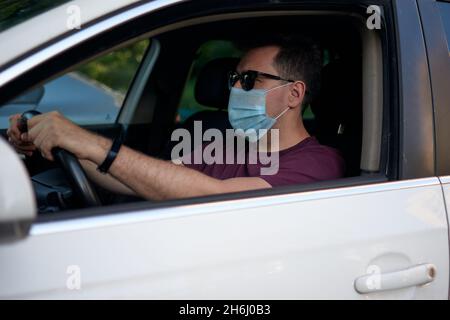 A man driving a car in a medical face mask during coronavirus outbreak, a taxi driver in a mask, protection from the virus. Driver in white car, hand Stock Photo