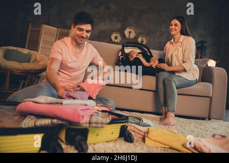 Photo of daddy put clothes luggage mommy sit sofa with daughter baby car seat ready for abroad weekend rest evening late indoors Stock Photo