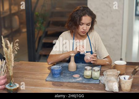 Young serious female painting handmade earthenware while sitting by table and holding one of three jars with paints Stock Photo