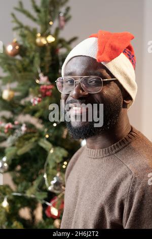Bearded young man in Santa cap and eyeglasses looking at you while standing against decorated Christmas tree Stock Photo