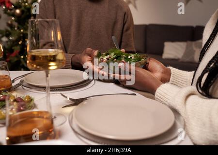 Hands of young African couple holding bowl with salad while sitting by served festive table and having dinner Stock Photo