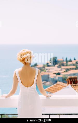 Bride in a white dress stands on a balcony looking at the island of Sveti Stefan near Budva, Montenegro. Back view Stock Photo