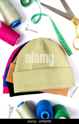 Top view of a stack of insulated stylish beanie hats in different colors with scissors, threads and needles. Tailoring concept. Vertical photo Stock Photo