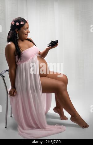 Studio photo of a happy Afro-American woman looking at some baby shoes. The woman is pregnant and her belly is visible Stock Photo