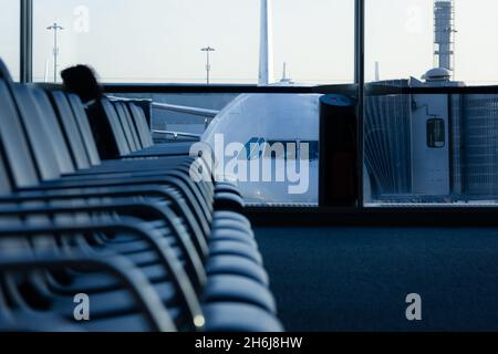 Empty row of seats at departure area with airplane connected to finger dock on background. Nobody at airport terminal. Futuristic, cold blue effect Stock Photo