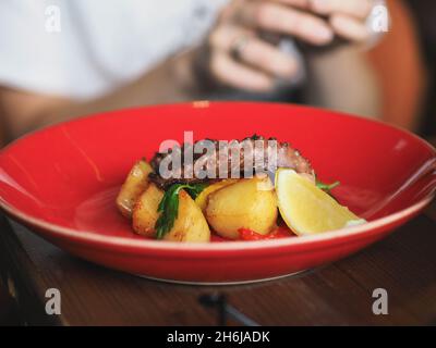 Fried octopus with potatoes on red plate in restaurant. Hands of young uncertain woman with cutlery. Front view Stock Photo