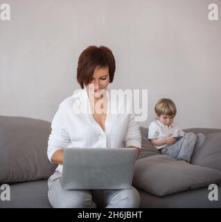 woman works on laptop at home, next to child is playing on phone. mom and son with digital gadgets are sitting together on sofa in living room. concep Stock Photo