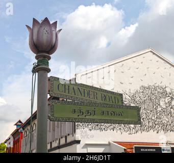 Green street name signs for Chander Road in English and Hindi with shophouses in the background and wall art in Little India, Singapore. Stock Photo