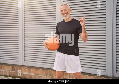 Happy sportive mature man in sportswear smiling at camera while standing outdoors with basketball ball Stock Photo