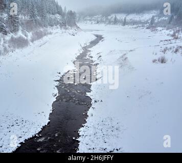 Narrow stream flows leisurely through the snowy landscape into the fog in the distance, monochrome Stock Photo