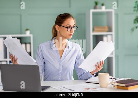 Focused businesswoman working on laptop and holding reports, sitting at workplace in office interior, reading documents Stock Photo