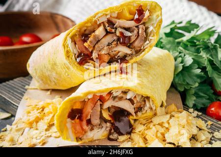 Crispy roll of shawarma with chips, barbeque sauce, chicken and vegetables in pita bread on wooden dark background with cherry tomatoes and green pars Stock Photo
