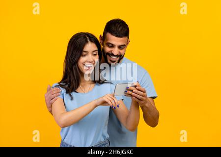 Joyful arab couple looking at smartphone screen, using new cool application, standing over yellow studio background Stock Photo