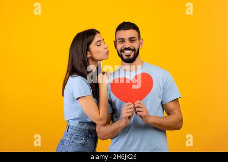 St. Valentines day concept. Portrait of loving arab couple holding red paper heart card, young woman kissing her husband Stock Photo