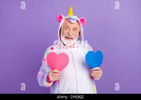 Portrait of attractive cheerful grey-haired man in kigurumi holding two hearts isolated over violet purple color background Stock Photo
