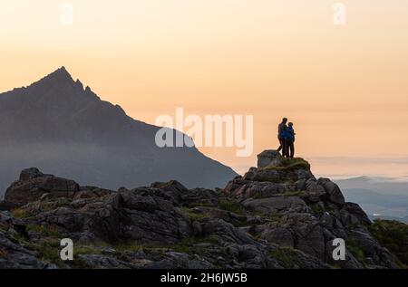 A couple standing on a rocky peak watching a mountain sunset with Sgurr nan Gillean in background, Isle of Skye, Inner Hebrides, Scotland Stock Photo