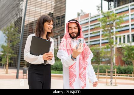 Smiling saudi arabia businessman wearing traditional clothes and young caucasian businesswoman using smartphone at meeting outdoors, arab man showing something on mobile phone to his female assistant Stock Photo