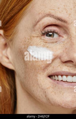Close up half face portrait of freckled red haired woman using face cream and smiling at camera Stock Photo