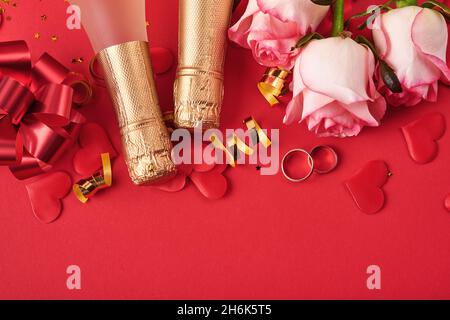 Pink roses flowers, wedding rings, champagne, gift, golden ribbons and confetti hearts on red background. Top view with space for greetings. Valentine Stock Photo