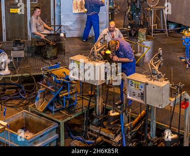 A worker at the Iittala glassworks, wearing blue dungarees, crosses the workshop floor; in the background, the opening of the melting furnace glows red-hot. At the moulding machine in the foreground, a glassmaker is working on a red-hot piece. The factory produces vases and figurines by hand and is now part of the Fiskars Group. Könnölänmäentie, Turkinmäki, Kalvola, Iittala, Hämeenlinna, Hämeenlinna sub-region, Kanta-Häme, Mainland Finland, 14500, Finland Stock Photo